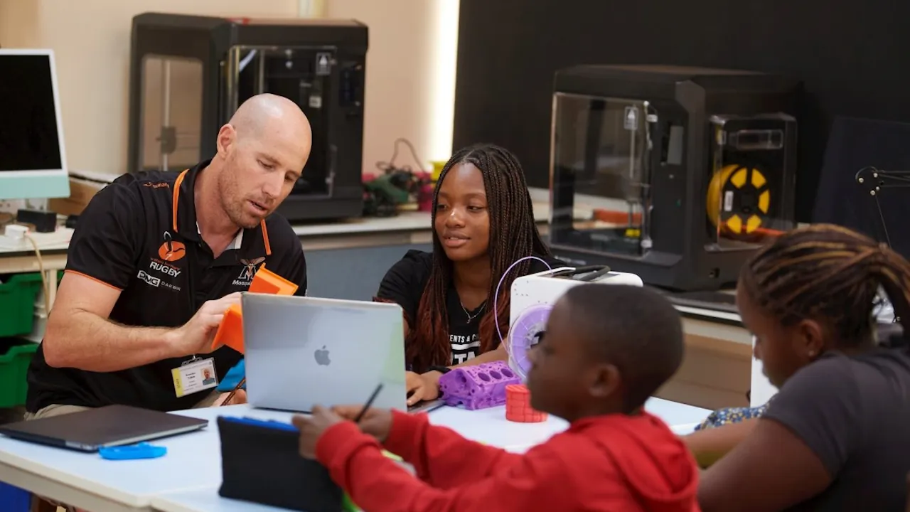 An instructor and students at the International School of Uganda engage with 3D printing technology and laptops in a modern design laboratory in Kampala.