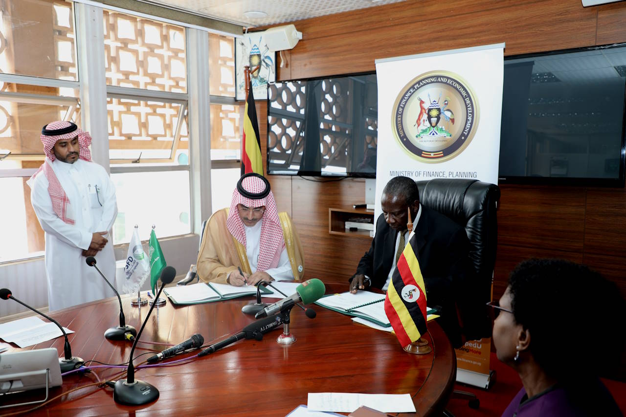 Two men, seated, in an office, sign documents as two other people, a female and a male, look on
